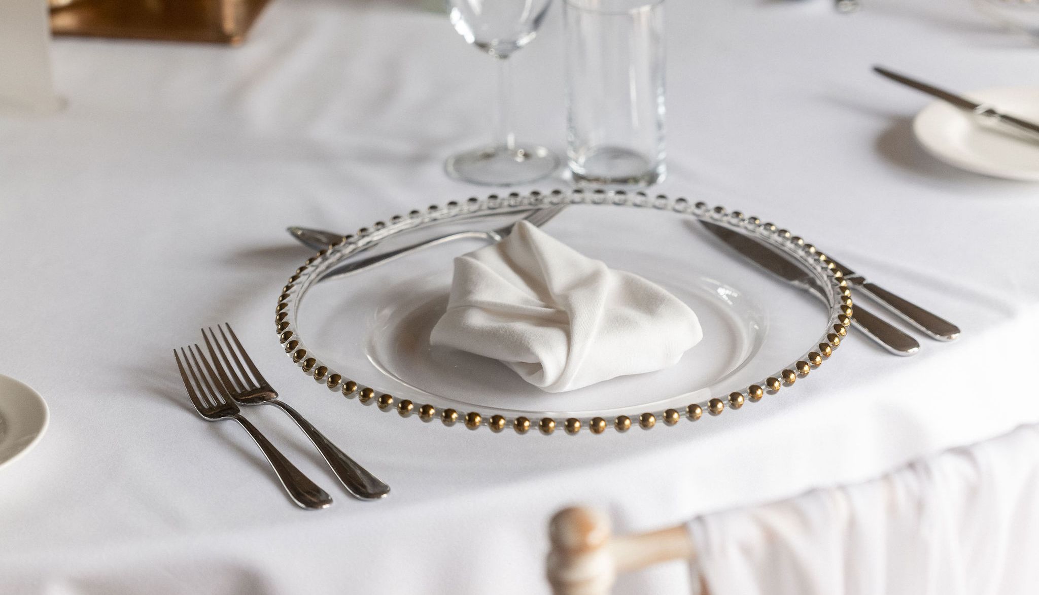 Elegant table setting with a white tablecloth, a decorative plate with a folded white napkin, silverware, glassware, and a small floral centerpiece.