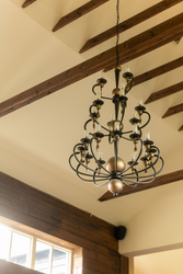 Elegant black and gold chandelier hanging from a ceiling with exposed wooden beams in a room with wood-paneled walls and high windows.