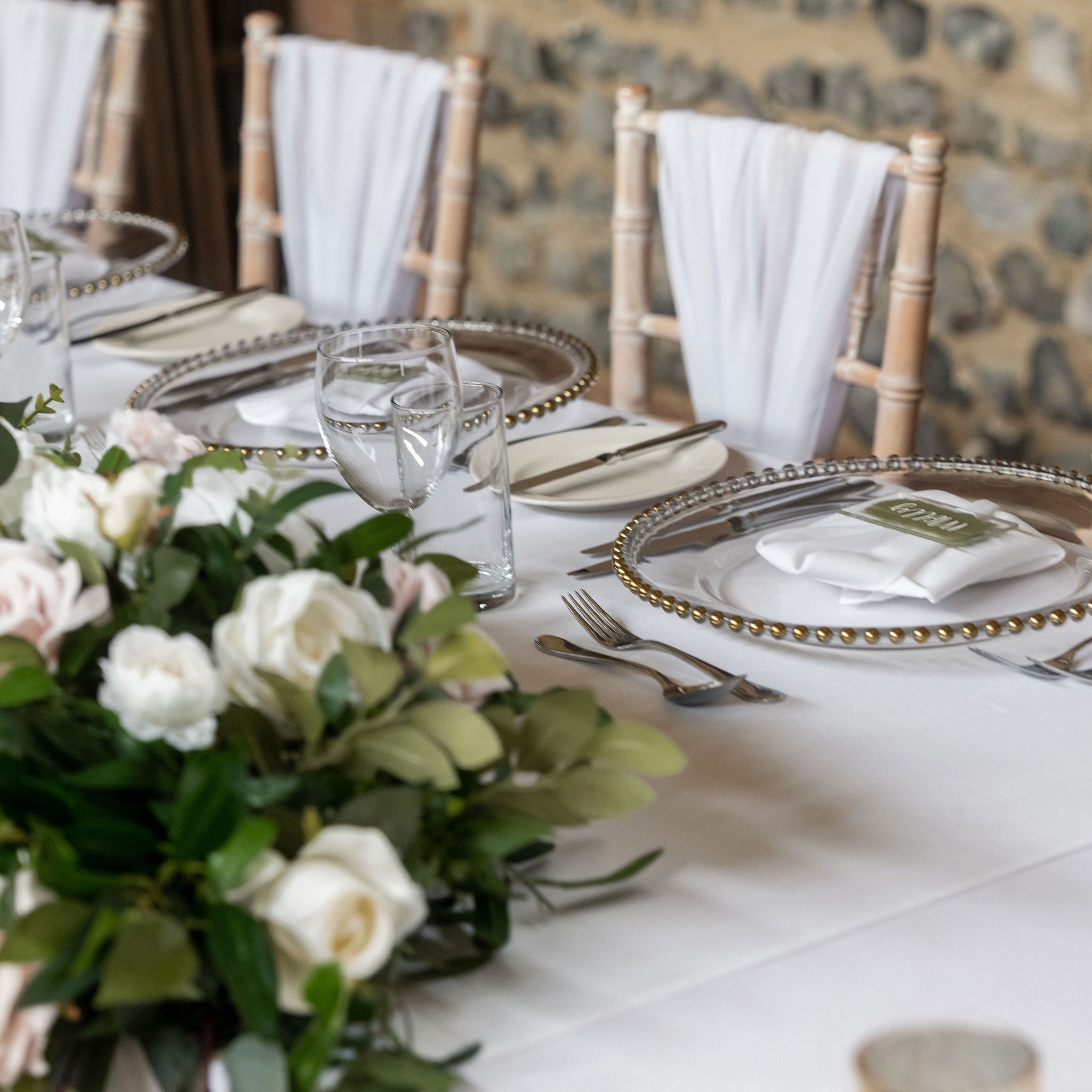 Elegant wedding or event table setting with white roses centerpiece, glassware, silver cutlery, decorative plates, and chairs draped with white fabric, set against a stone wall background.