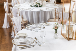 Elegant wedding reception table setting with white linens, gold-rimmed plates, glassware, floral centerpieces, and gold chairs draped with white fabric.