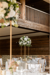 Elegant wedding reception setup with tall floral centerpieces, white tablecloths, wooden chairs, glassware, and a rustic wooden interior.