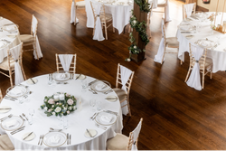 Elegant wedding reception setup with round tables covered in white tablecloths, gold rimmed plates, flower centerpieces, and chairs draped with white fabric on a wooden floor.