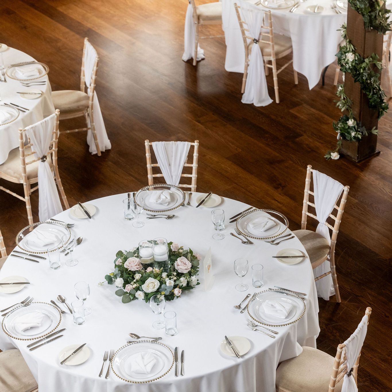Elegant wedding reception setup with round tables covered in white tablecloths, gold rimmed plates, flower centerpieces, and chairs draped with white fabric on a wooden floor.