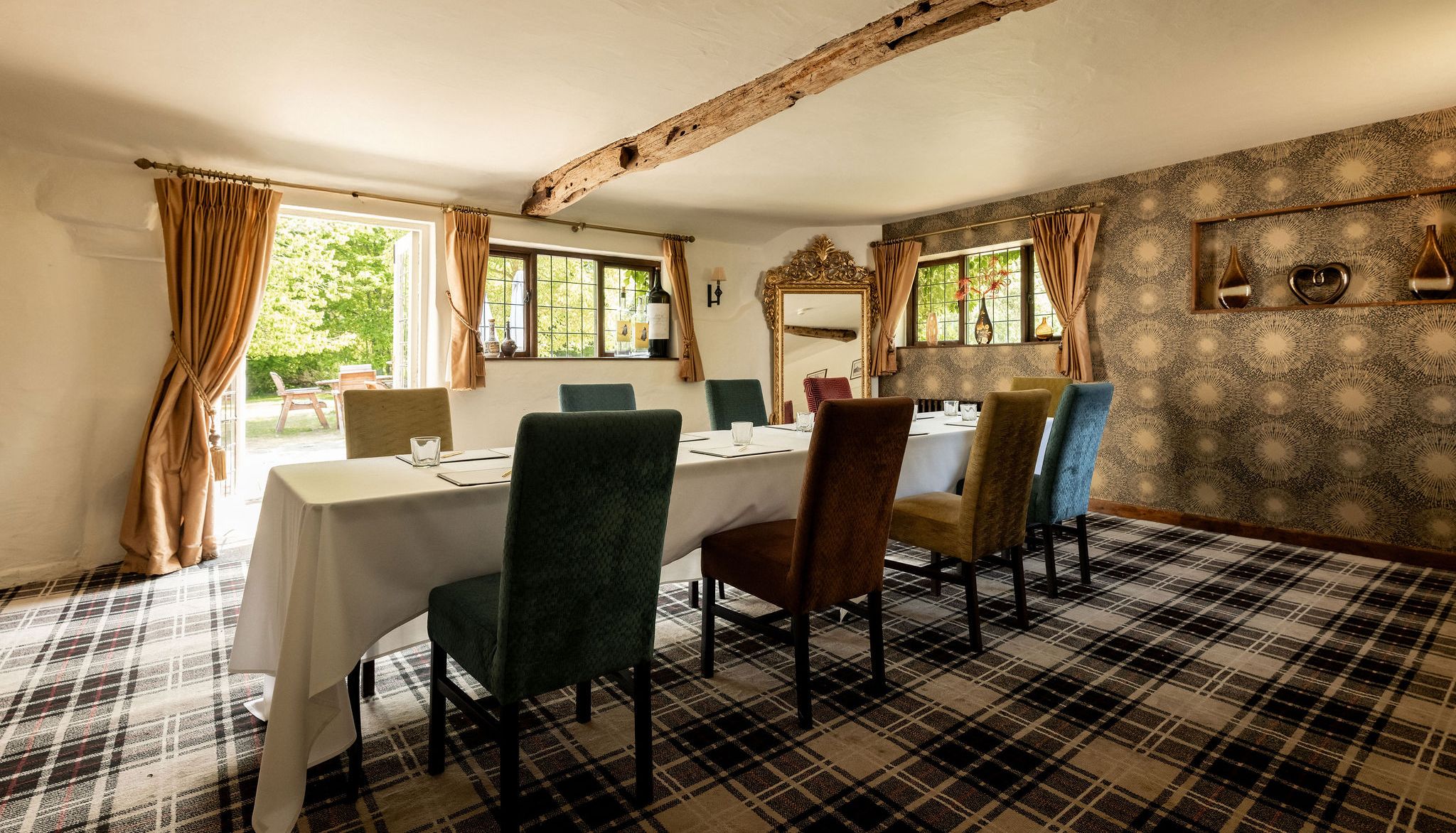 Elegant dining or meeting room with a set table and colorful chairs, featuring rustic wooden beams, patterned wallpaper, and large windows with gold curtains overlooking a garden.