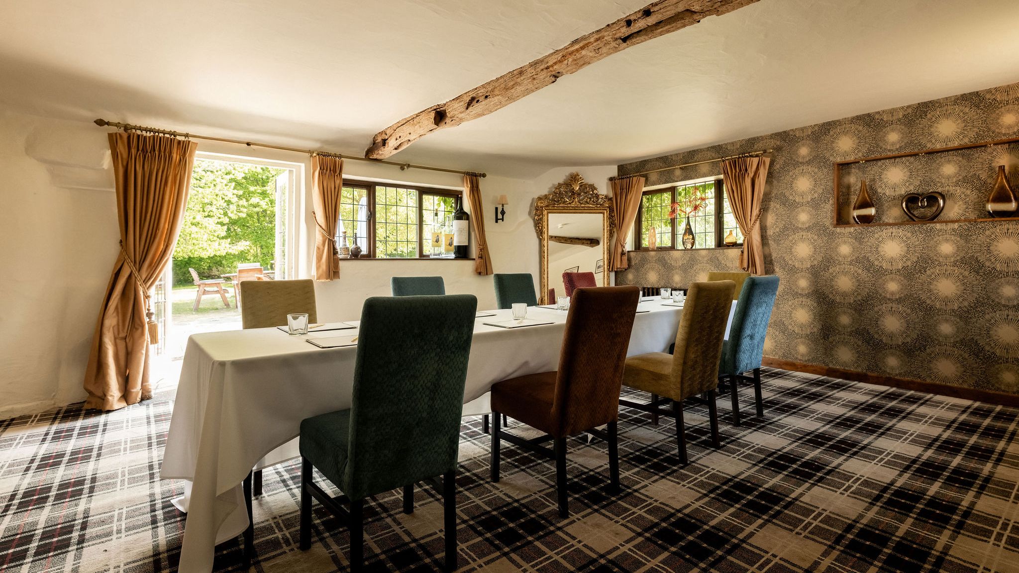 Elegant dining or meeting room with a set table and colorful chairs, featuring rustic wooden beams, patterned wallpaper, and large windows with gold curtains overlooking a garden.