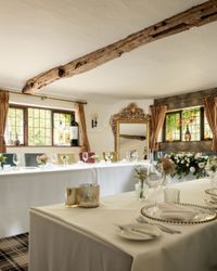 Elegant dining room with rustic wooden beam, floral arrangements, and neatly set tables with glassware and gold-rimmed plates.