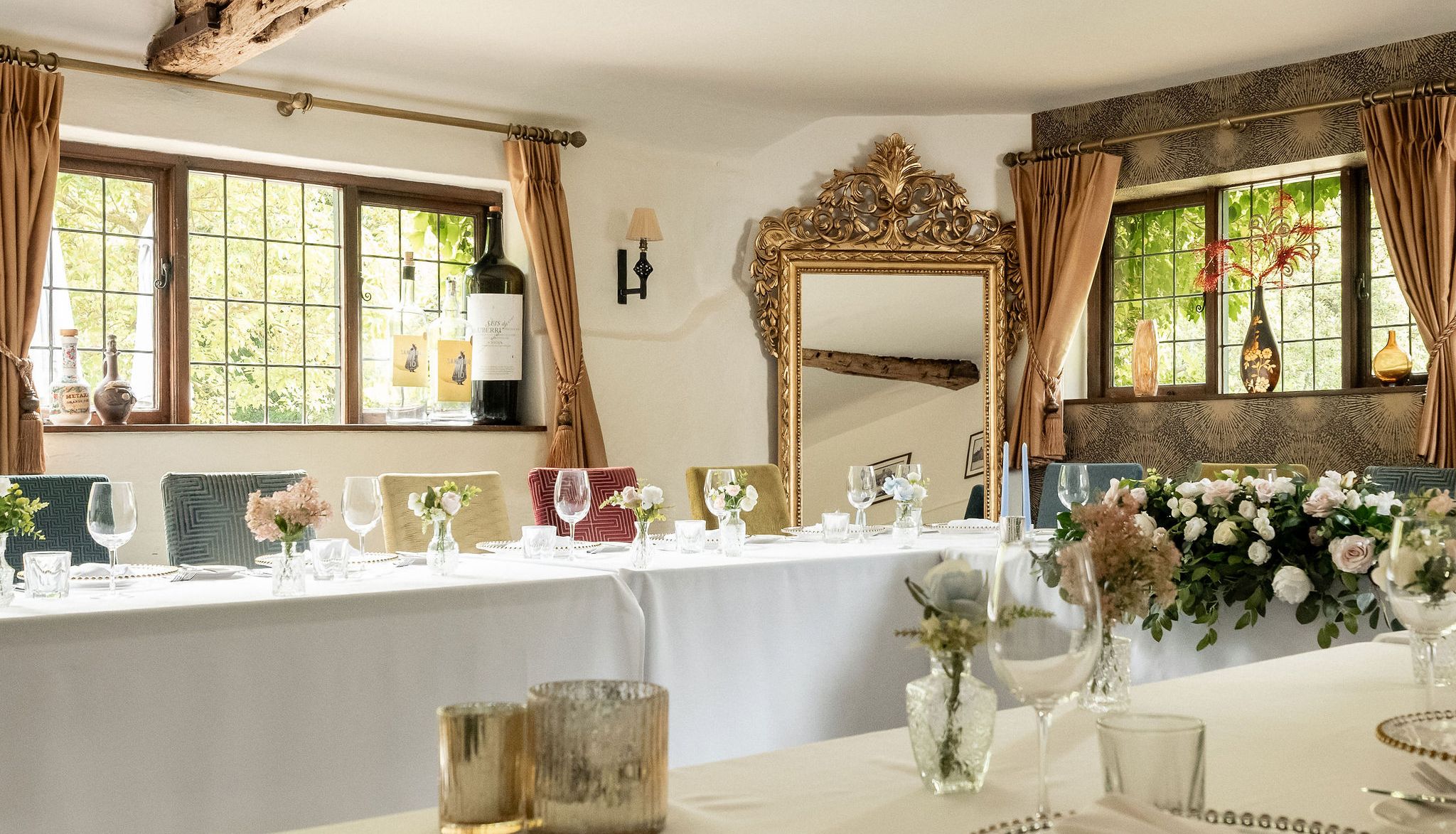 Elegant dining room with rustic wooden beam, floral arrangements, and neatly set tables with glassware and gold-rimmed plates.