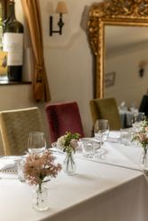 Elegant dining room setup with a white tablecloth, glassware, small floral vases, and upholstered chairs beside a large ornate mirror.