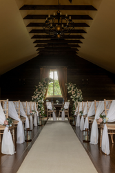 Rustic barn wedding ceremony setup with wooden chairs draped in white fabric, aisle lined with lanterns, and a floral arch by a window.