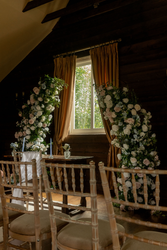 Indoor wedding ceremony setup in a rustic barn with floral arch decorations and wooden chairs facing a table by a window.