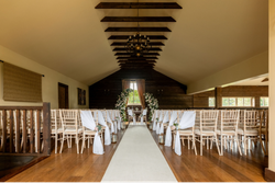 Elegant wedding ceremony setup in a rustic barn with wooden chairs, white drapery, and floral arrangements leading to an arch in front of a window.
