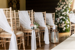 Rows of wooden chairs with white drapes and floral decorations set up for a wedding ceremony in a barn.