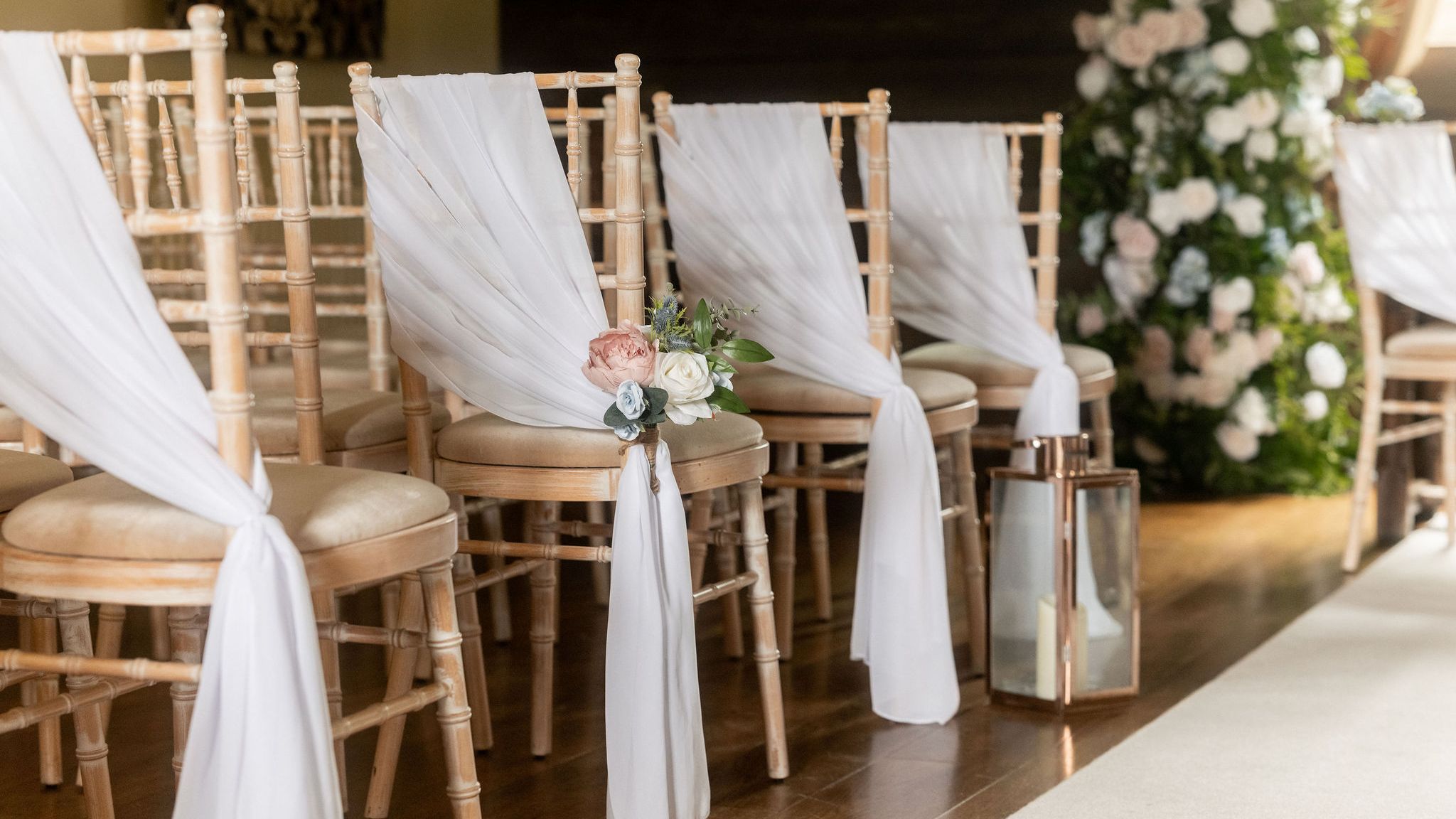 Rows of wooden chairs with white drapes and floral decorations set up for a wedding ceremony in a barn.