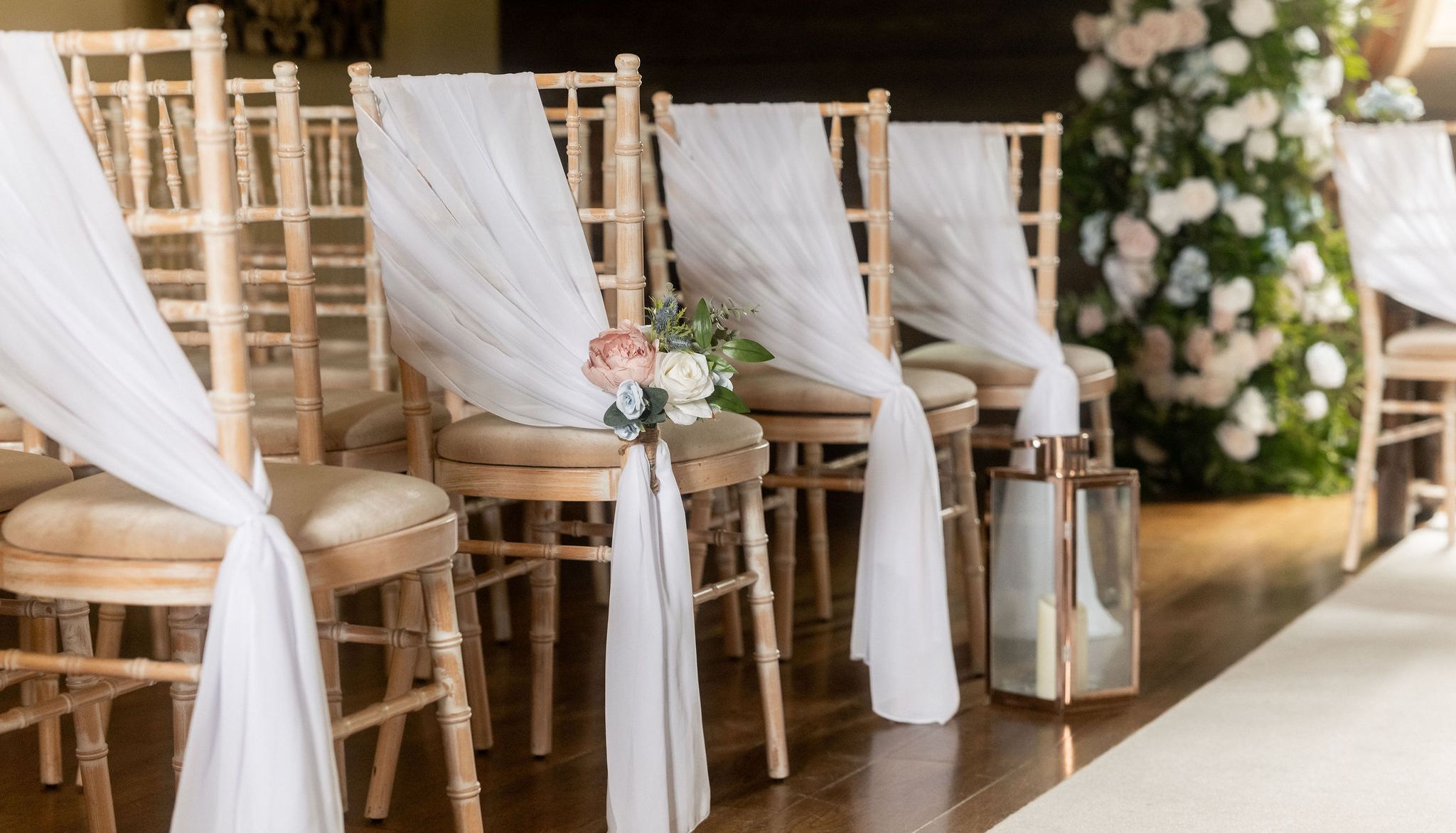 Rows of wooden chairs with white drapes and floral decorations set up for a wedding ceremony in a barn.