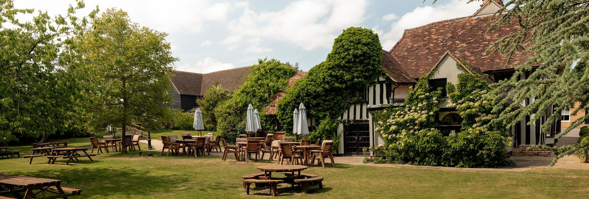 Outdoor seating area with wooden tables and chairs on a grassy lawn in front of a rustic building covered in green ivy.