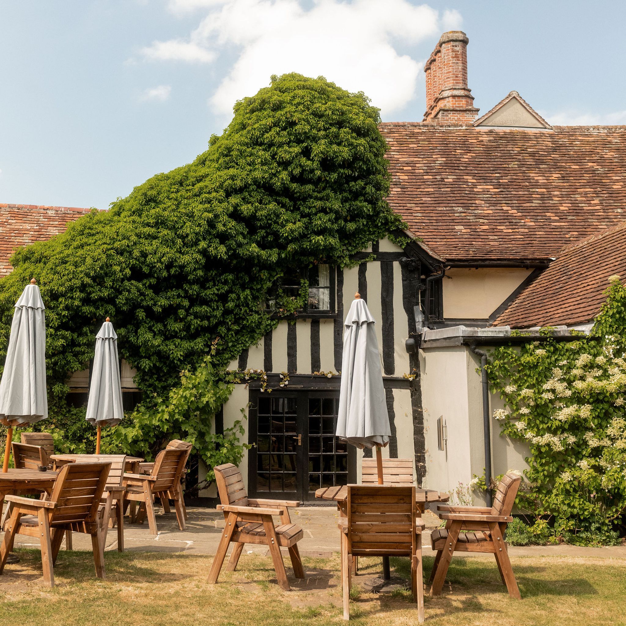 Outdoor seating area with wooden tables and chairs in front of a traditional building covered in green ivy and plants, with large windows and a tiled roof.