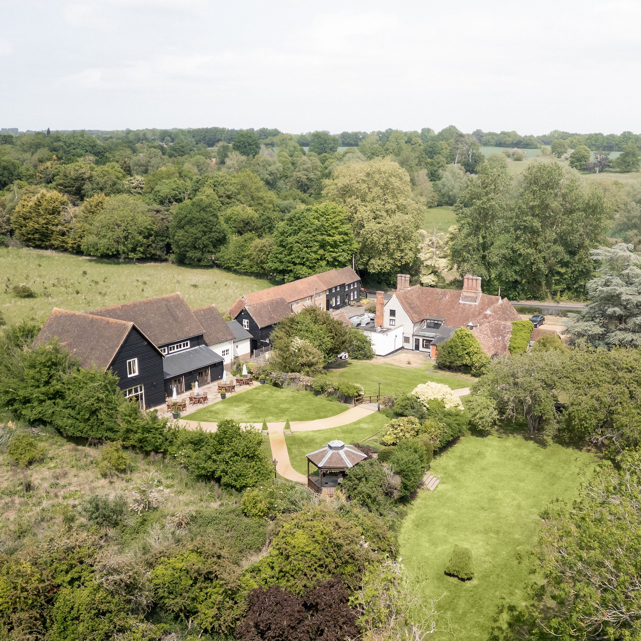 Aerial view of a countryside estate with several buildings, a gazebo, gardens, and large grassy areas surrounded by trees.