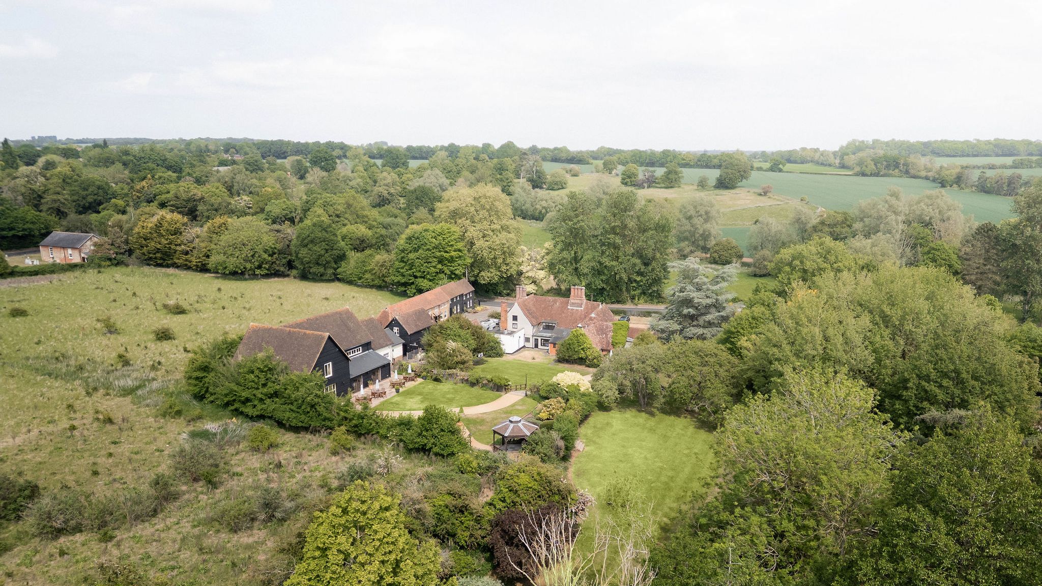 Aerial view of a countryside estate with multiple buildings and lush green gardens surrounded by fields and trees.