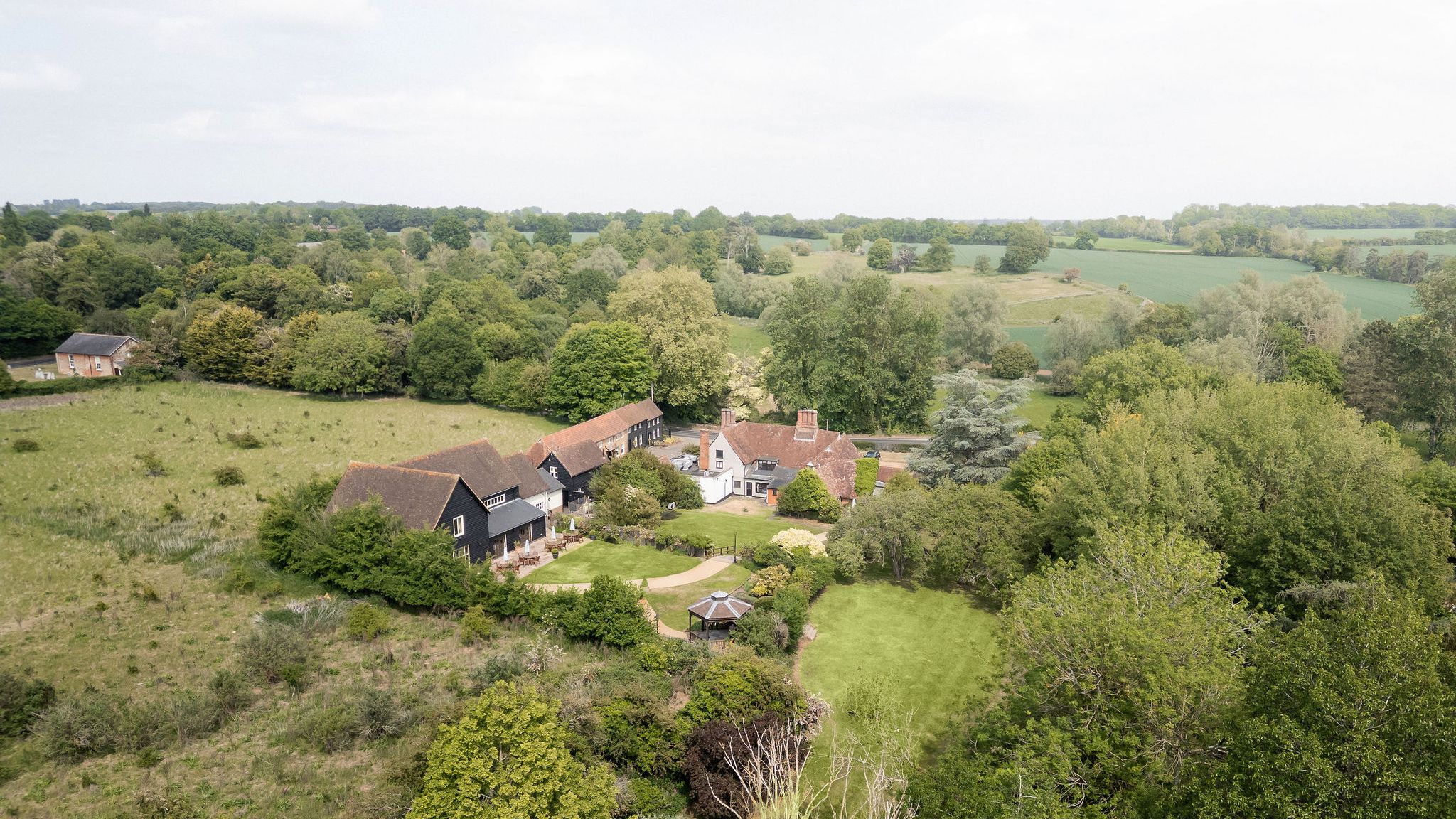 Aerial view of a countryside estate with multiple buildings and lush green gardens surrounded by fields and trees.