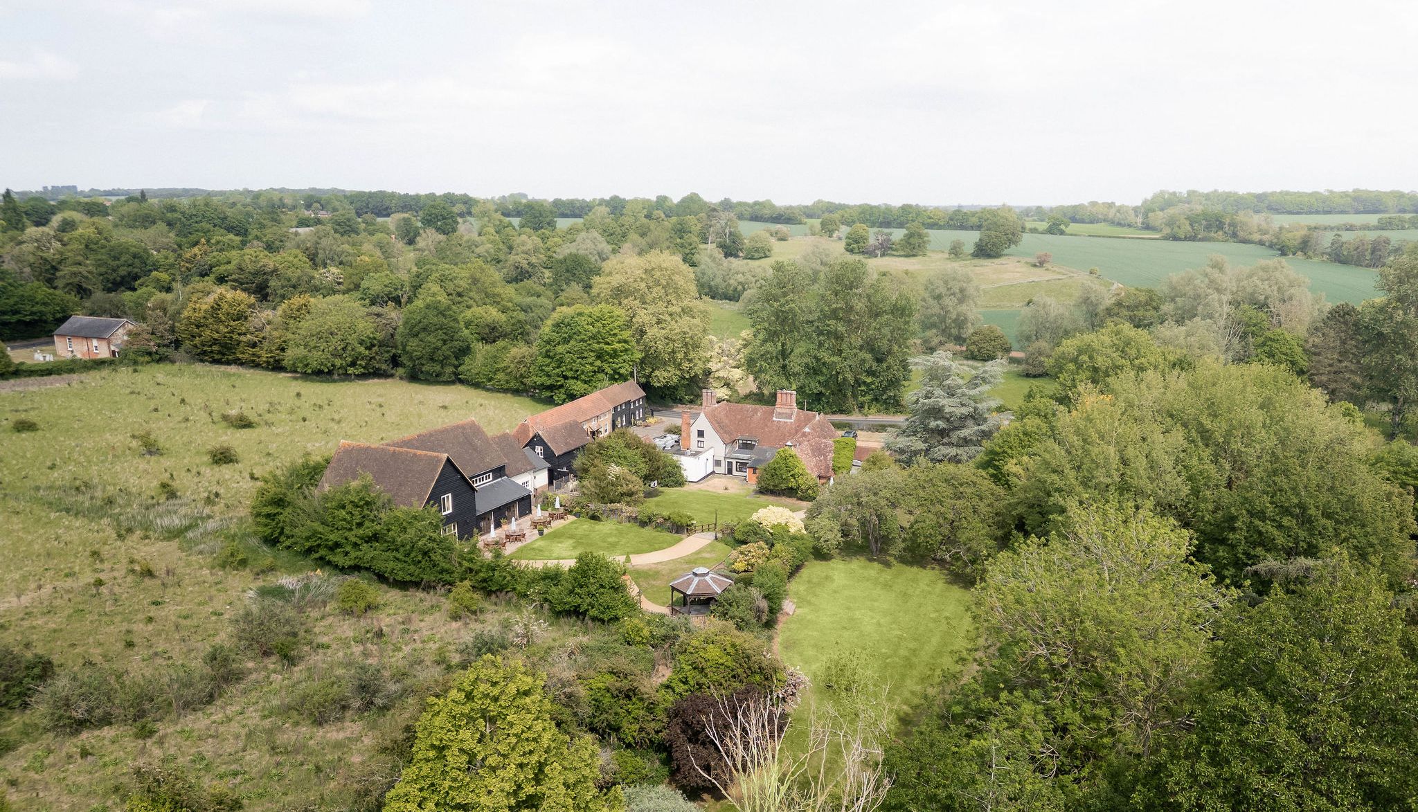 Aerial view of a countryside estate with multiple buildings and lush green gardens surrounded by fields and trees.