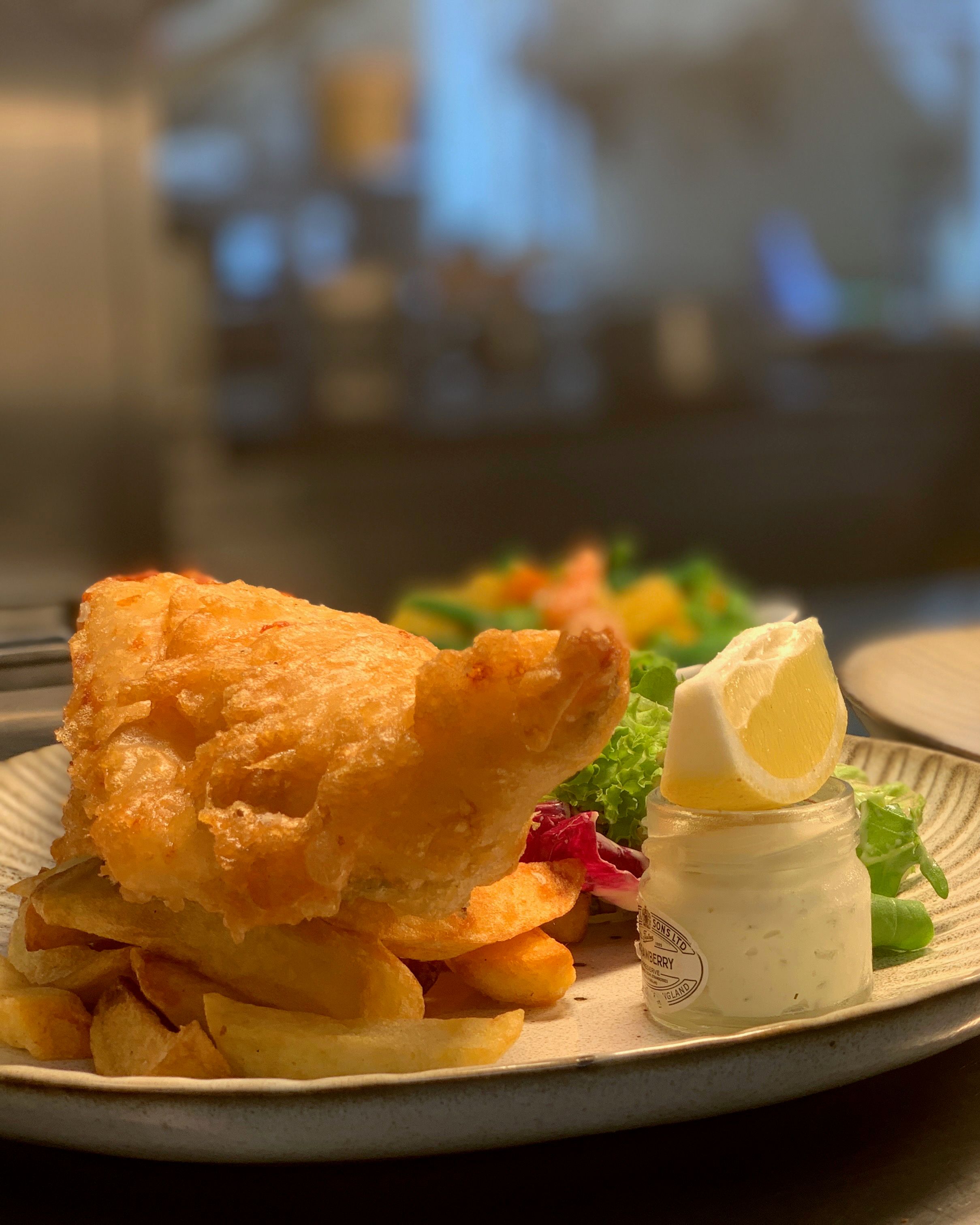 A plate of fish and chips with a jar of tartar sauce and lemon wedge, alongside another plate with layered potatoes and meat in the background.