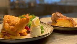 A plate of fish and chips with a jar of tartar sauce and lemon wedge, alongside another plate with layered potatoes and meat in the background.