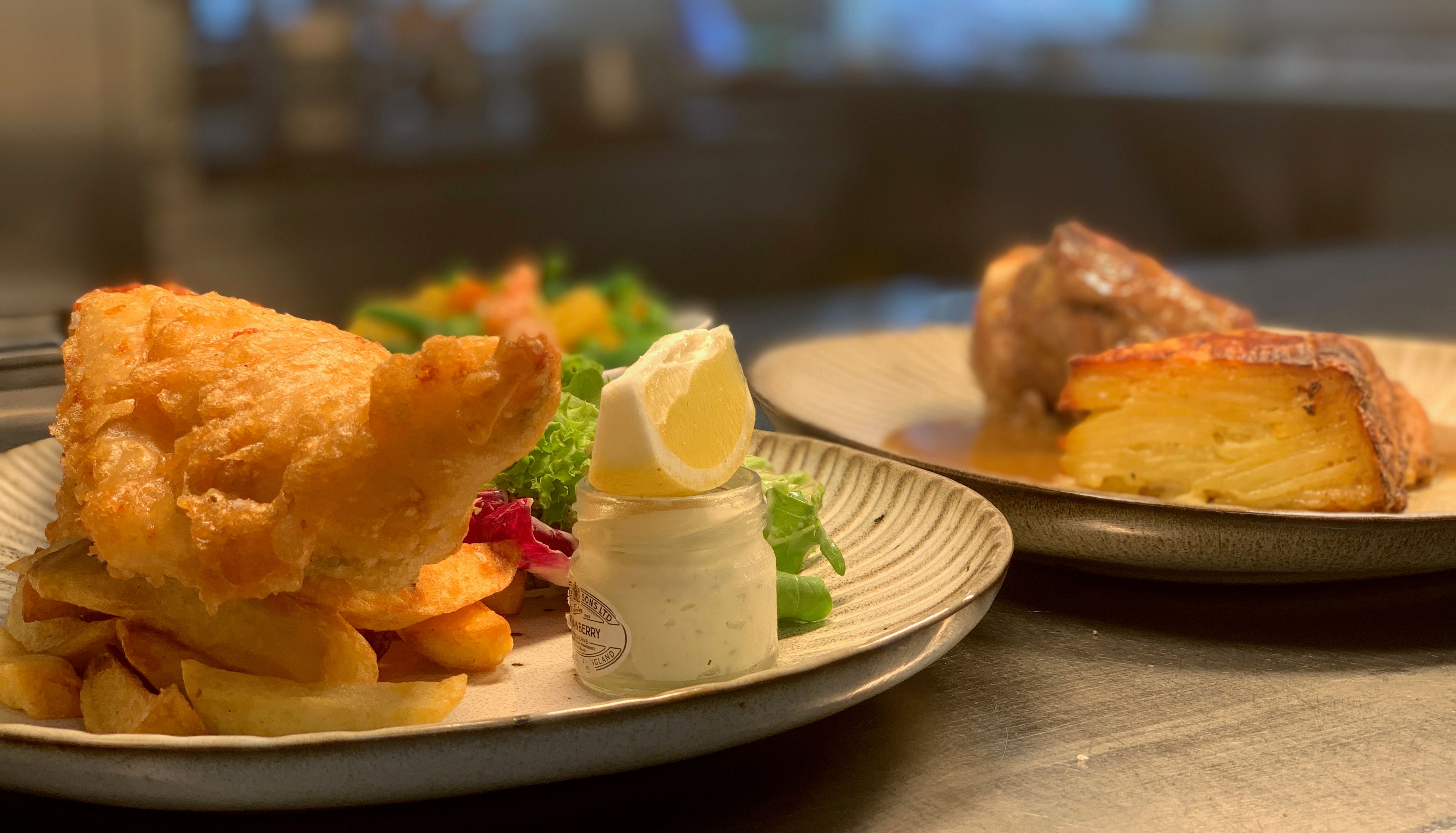 A plate of fish and chips with a jar of tartar sauce and lemon wedge, alongside another plate with layered potatoes and meat in the background.