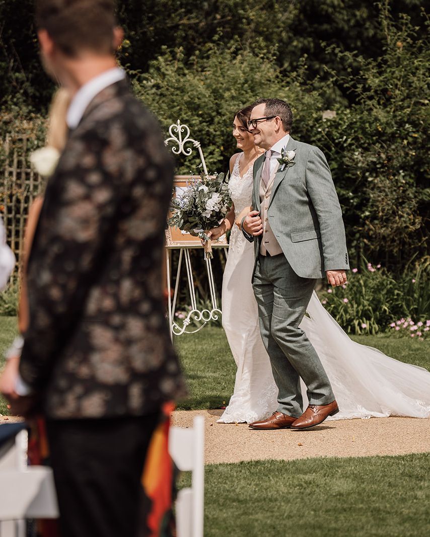 Bride and a man walking down an outdoor aisle at a wedding with guests in the foreground.