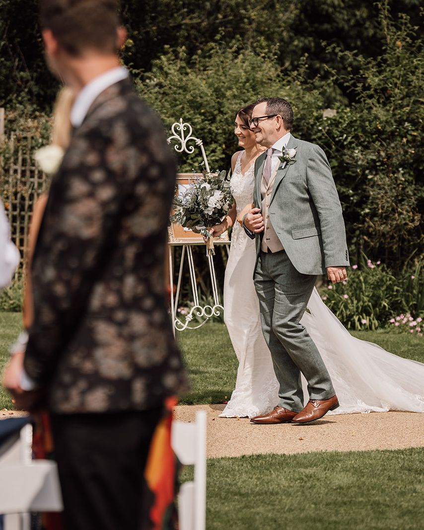 Bride and a man walking down an outdoor aisle at a wedding with guests in the foreground.