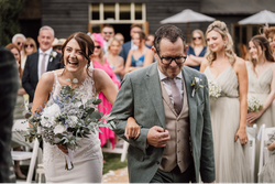 Bride laughing as she walks down the aisle arm-in-arm with a man in a suit, carrying a bouquet, with bridesmaids and guests in the background.