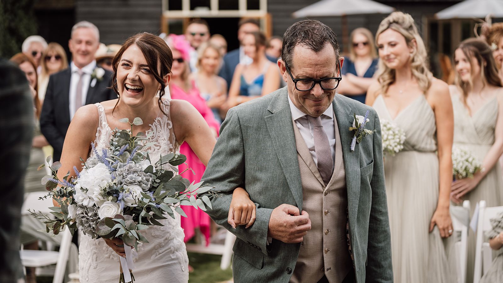 Bride laughing as she walks down the aisle arm-in-arm with a man in a suit, carrying a bouquet, with bridesmaids and guests in the background.