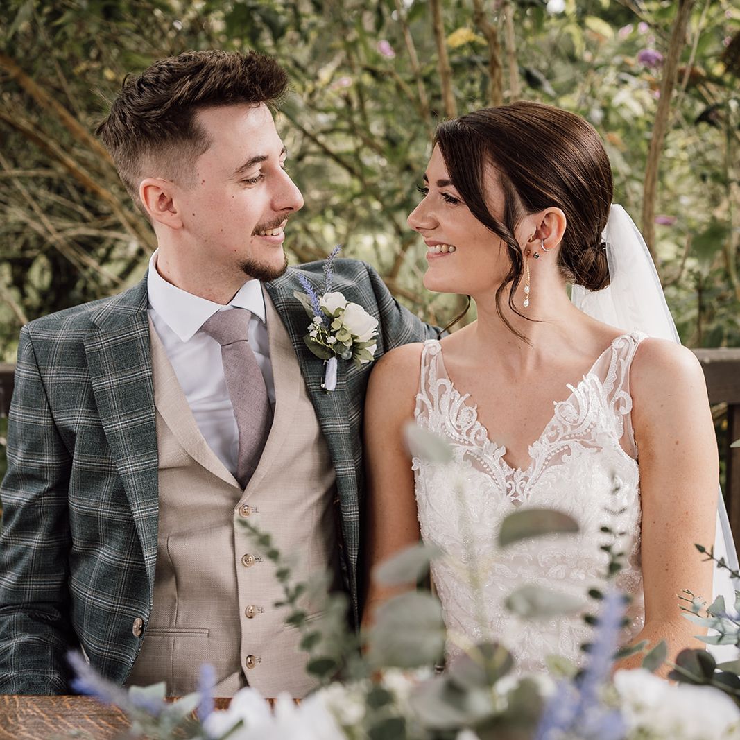 Smiling couple dressed in wedding attire sitting together outdoors among greenery and flowers.