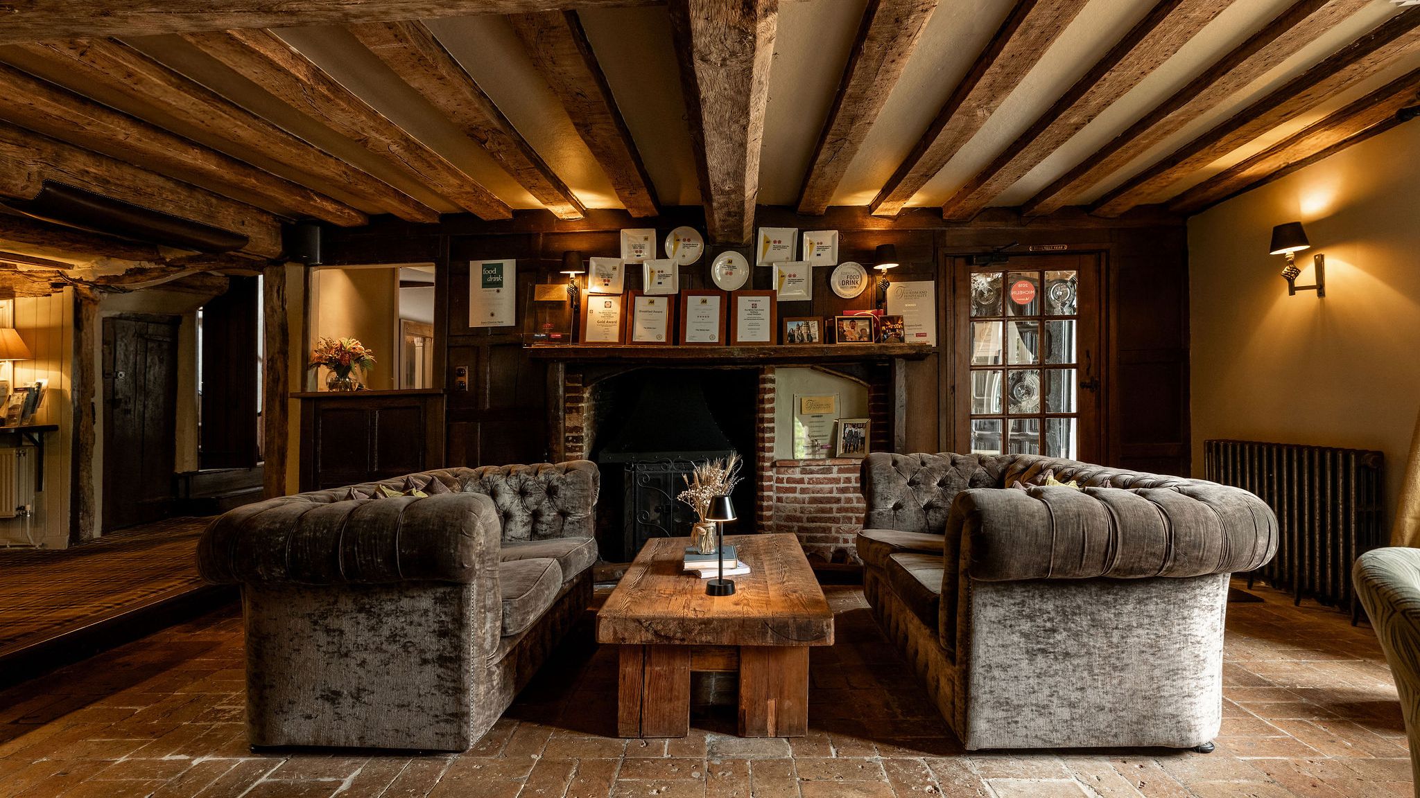 Cozy rustic lounge area with two velvet sofas facing each other, a wooden coffee table, exposed wooden beams, and a fireplace adorned with awards.