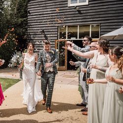 Bride and groom walking hand in hand while guests throw flower petals at their wedding celebration outside a rustic building.