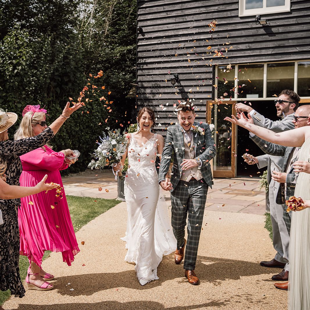 Bride and groom walking hand in hand while guests throw flower petals at their wedding celebration outside a rustic building.