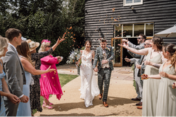 Bride and groom walking hand in hand while guests throw flower petals at their wedding celebration outside a rustic building.