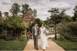 Bride and groom walking outside on a path, smiling at each other, with a garden and historic building in the background.