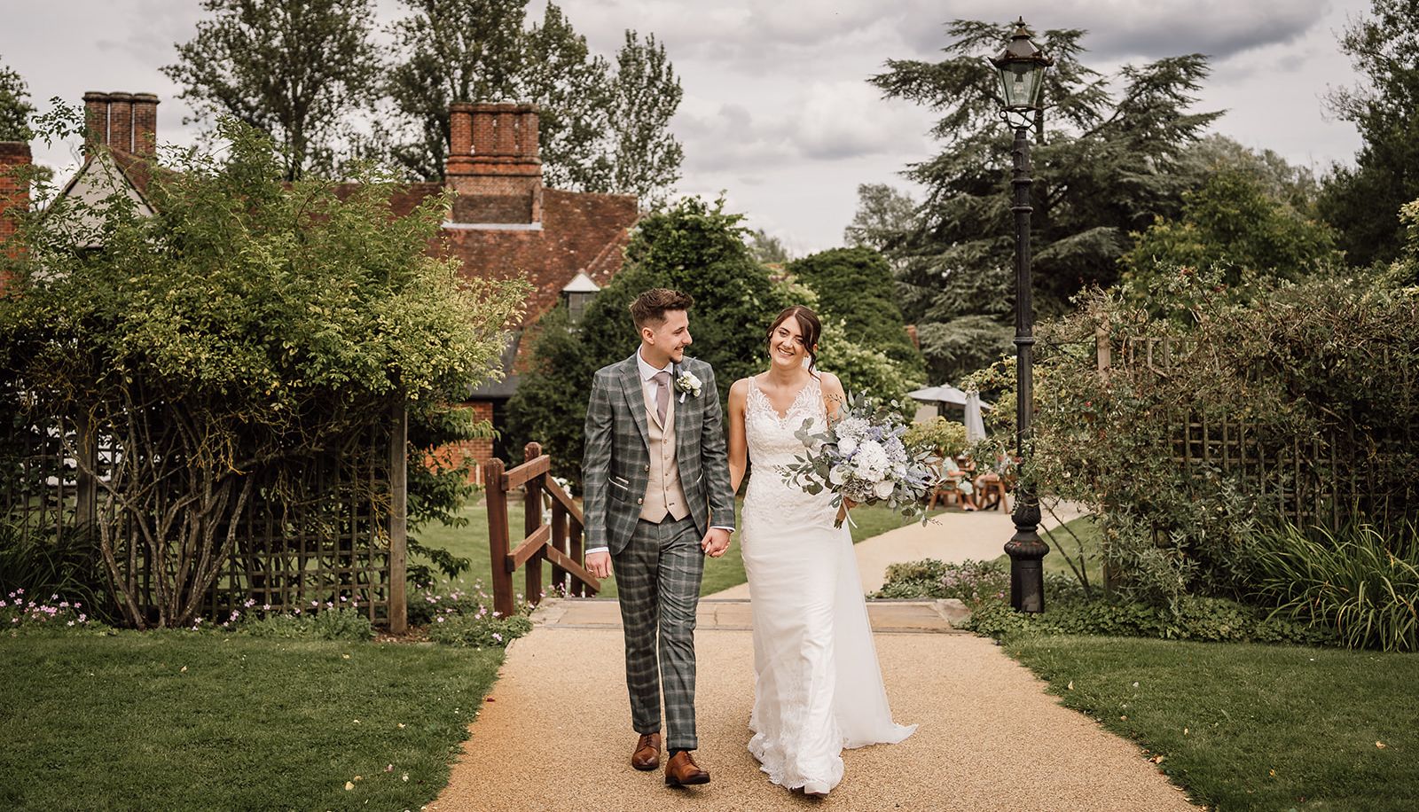 Bride and groom walking outside on a path, smiling at each other, with a garden and historic building in the background.