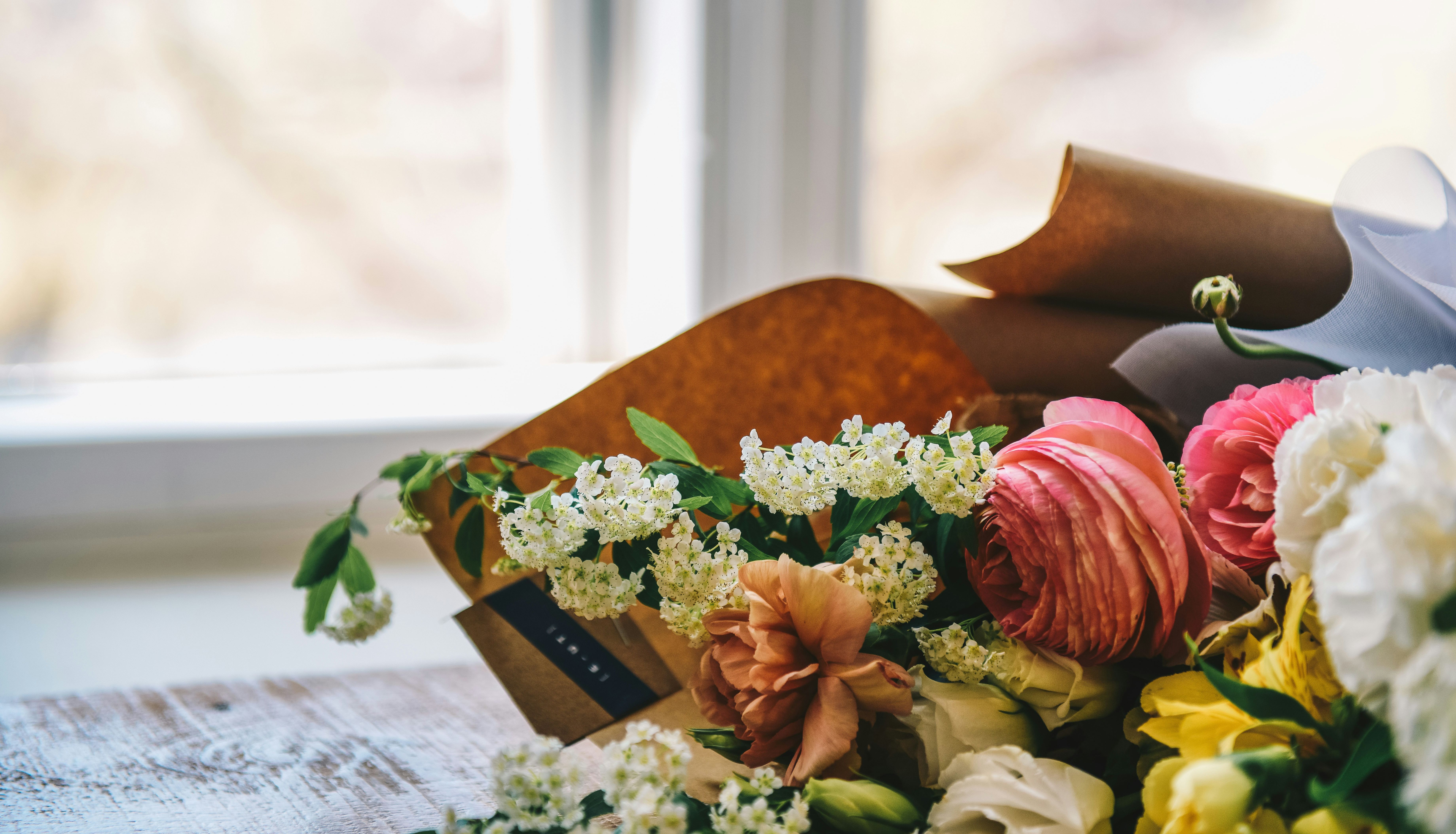 A close-up of a colorful bouquet of flowers, including pink ranunculus and white blossoms, placed on a wooden table near a window.