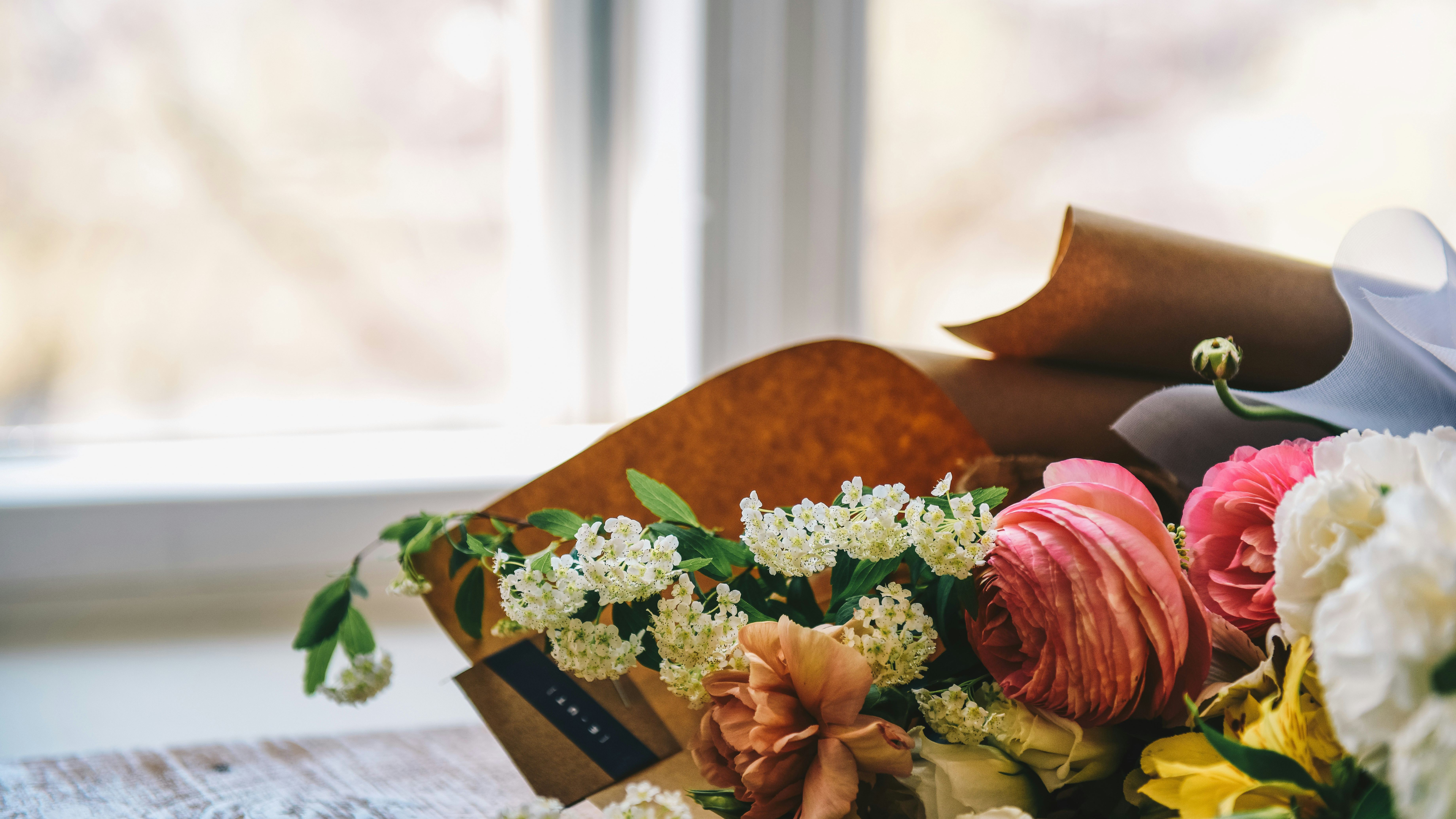 A close-up of a colorful bouquet of flowers, including pink ranunculus and white blossoms, placed on a wooden table near a window.