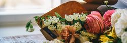 A close-up of a colorful bouquet of flowers, including pink ranunculus and white blossoms, placed on a wooden table near a window.