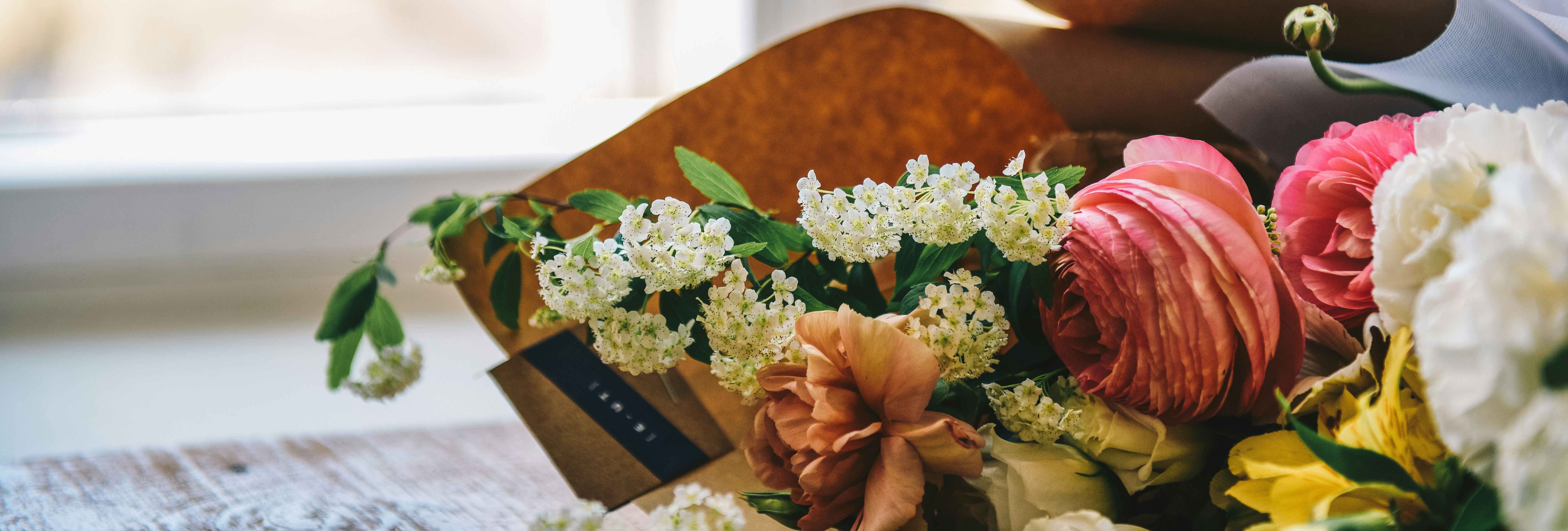 A close-up of a colorful bouquet of flowers, including pink ranunculus and white blossoms, placed on a wooden table near a window.