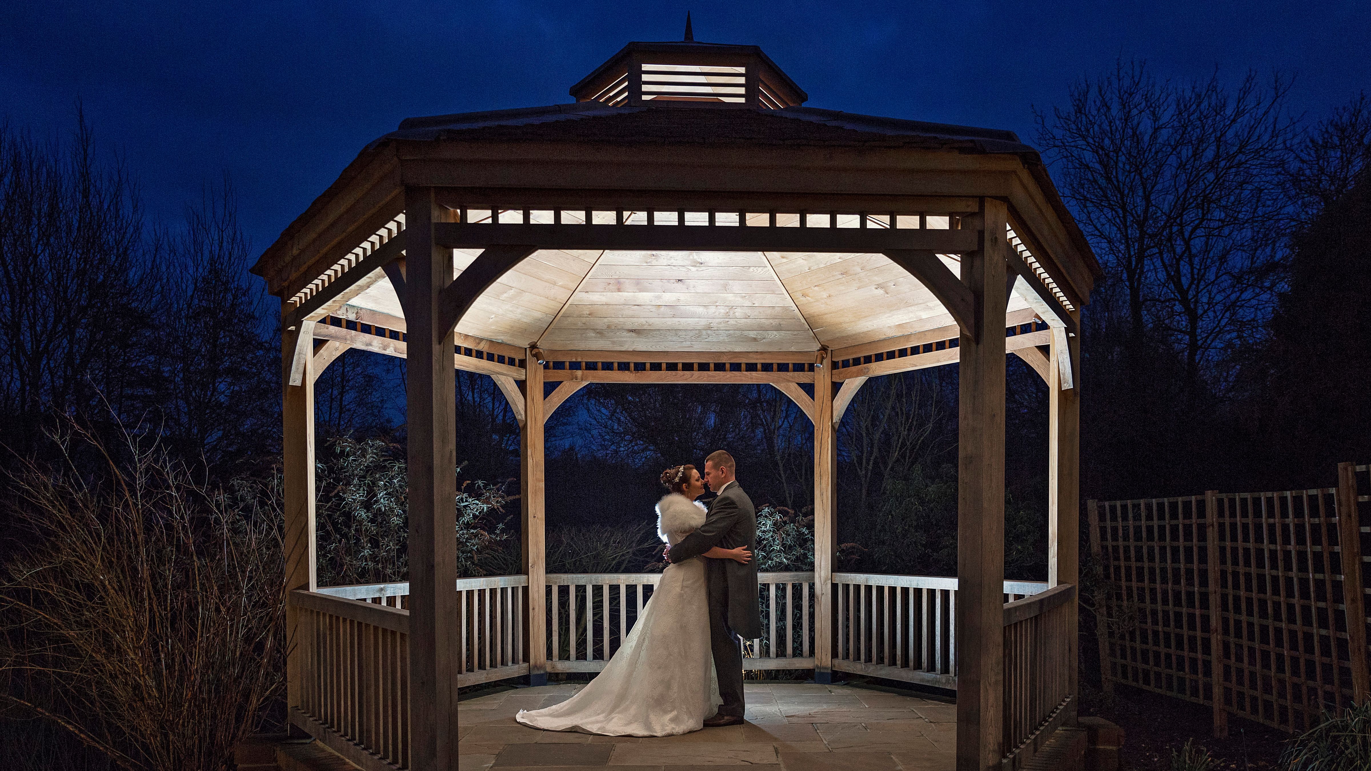 Bride and groom embrace in a beautifully lit wooden gazebo at night.