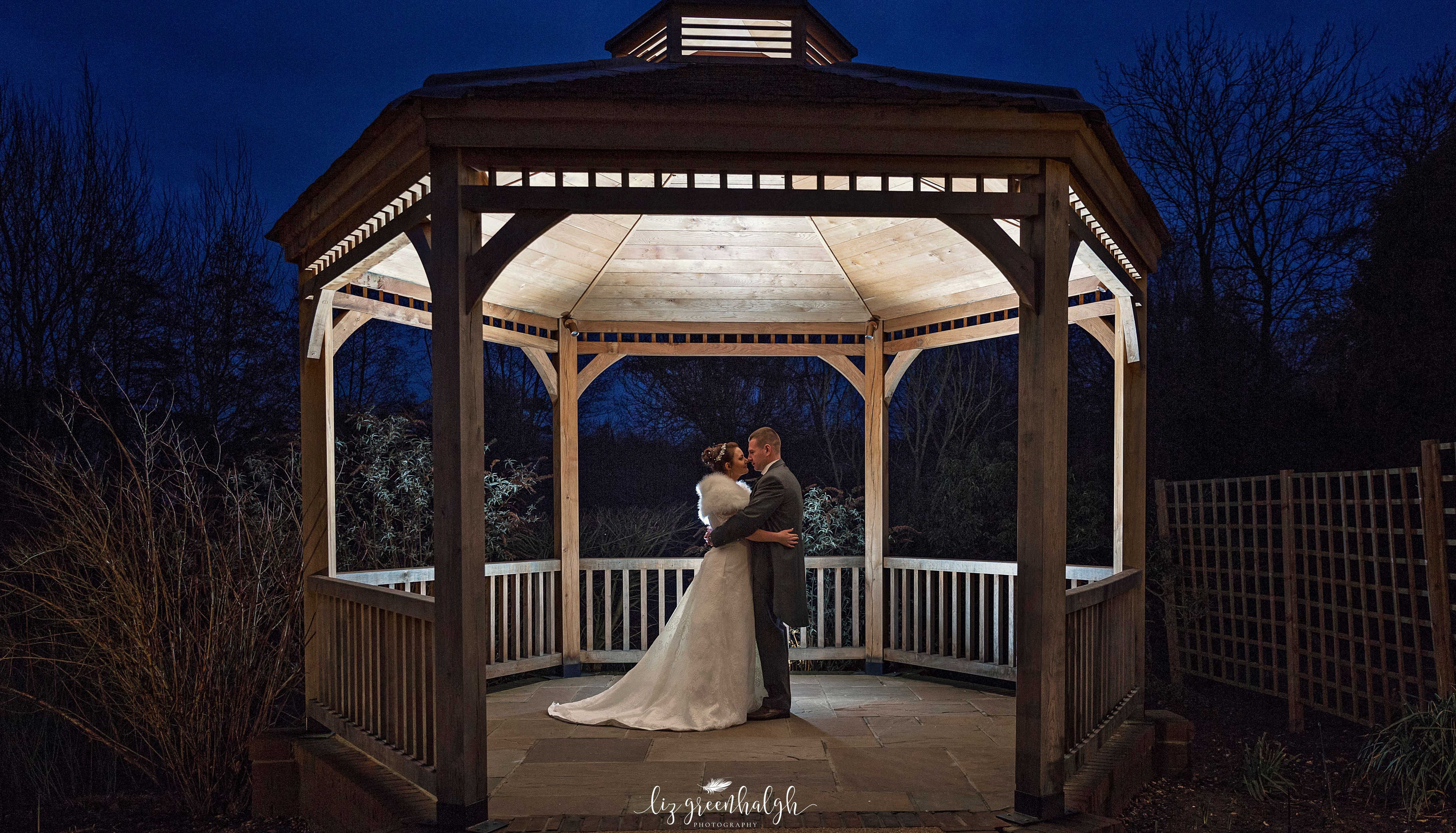 Bride and groom embrace in a beautifully lit wooden gazebo at night.