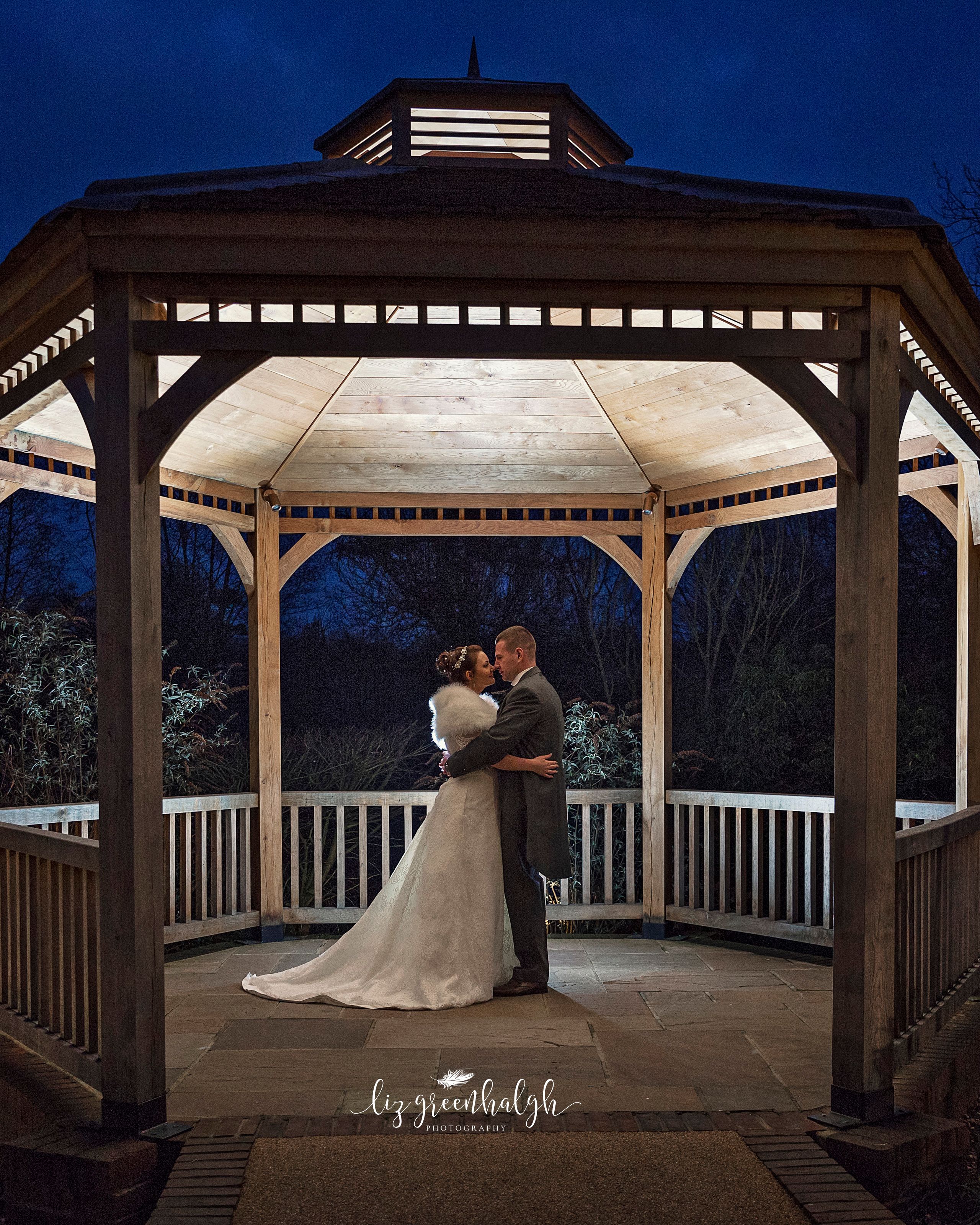 Bride and groom embrace in a beautifully lit wooden gazebo at night.