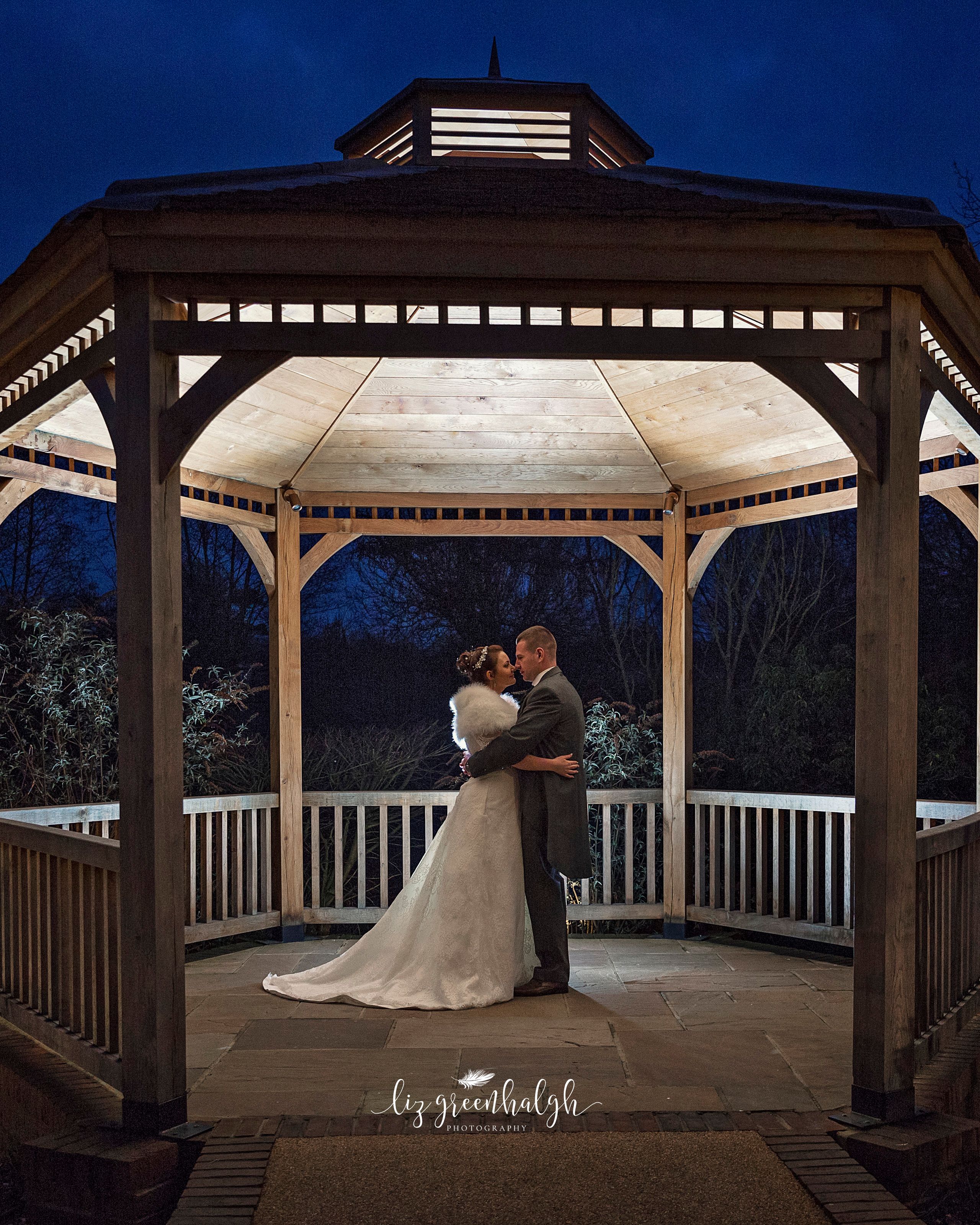 Bride and groom embrace in a beautifully lit wooden gazebo at night.