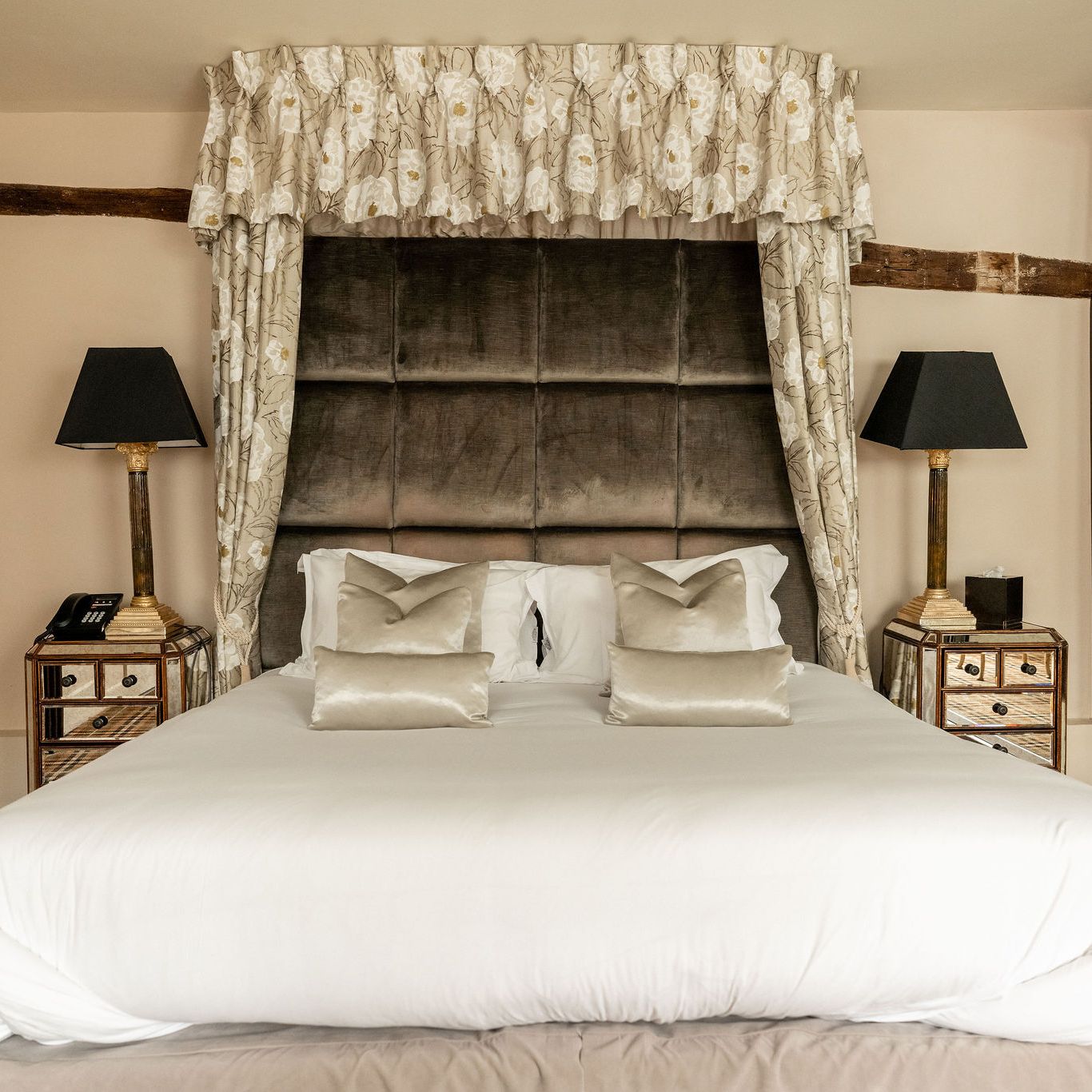 Luxurious bedroom with a large upholstered headboard, decorative floral canopy, white bedding, mirrored nightstands with lamps, and a plaid carpet.