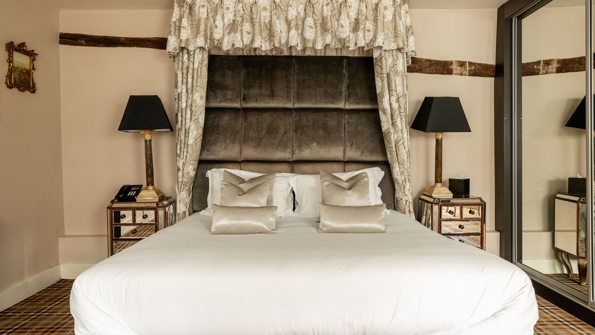 Luxurious bedroom with a large upholstered headboard, decorative floral canopy, white bedding, mirrored nightstands with lamps, and a plaid carpet.