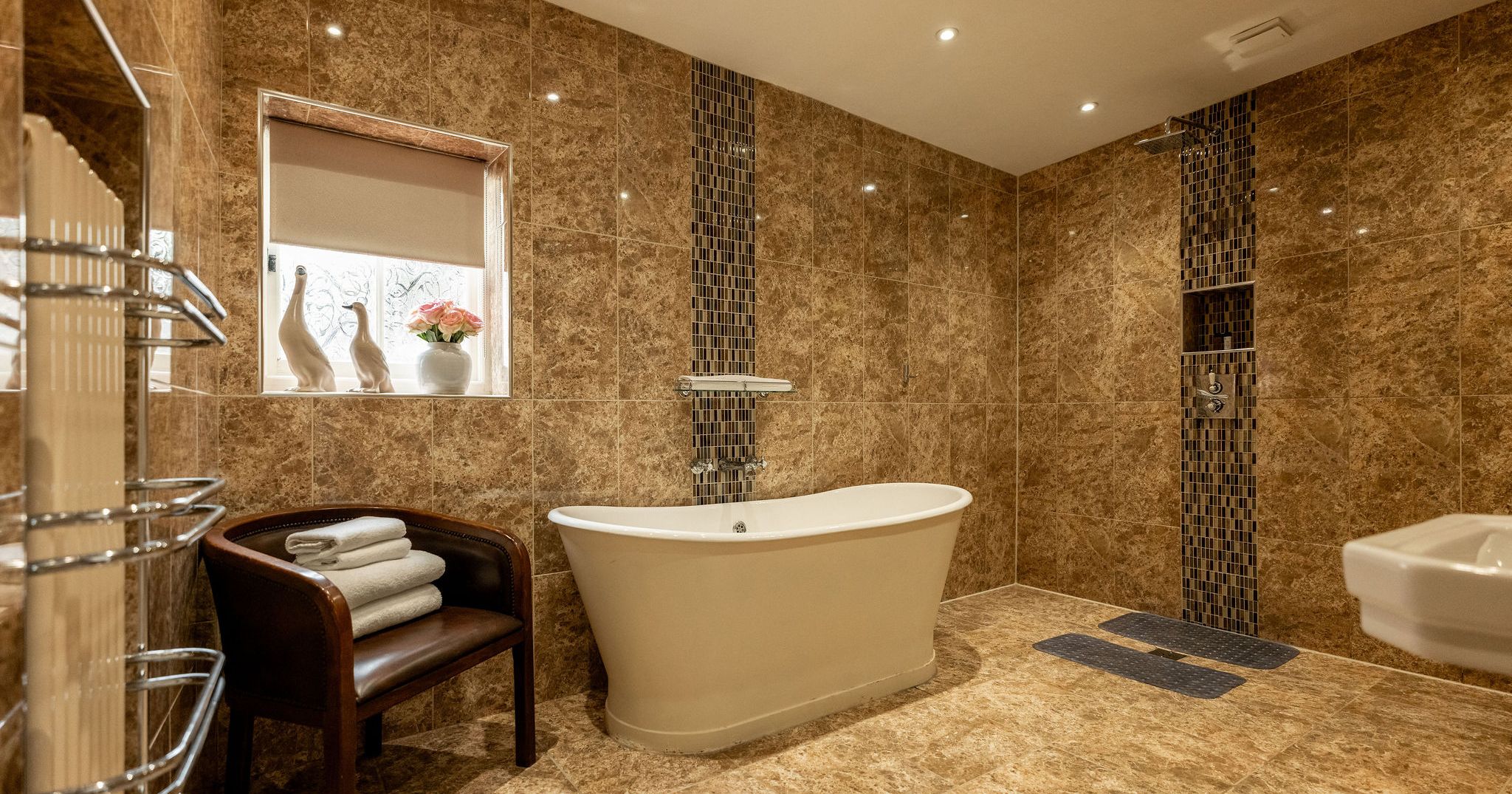 Luxurious bathroom with brown marble tiles, freestanding bathtub, window with decorative vases and flowers, a chair with folded towels, and modern fixtures.