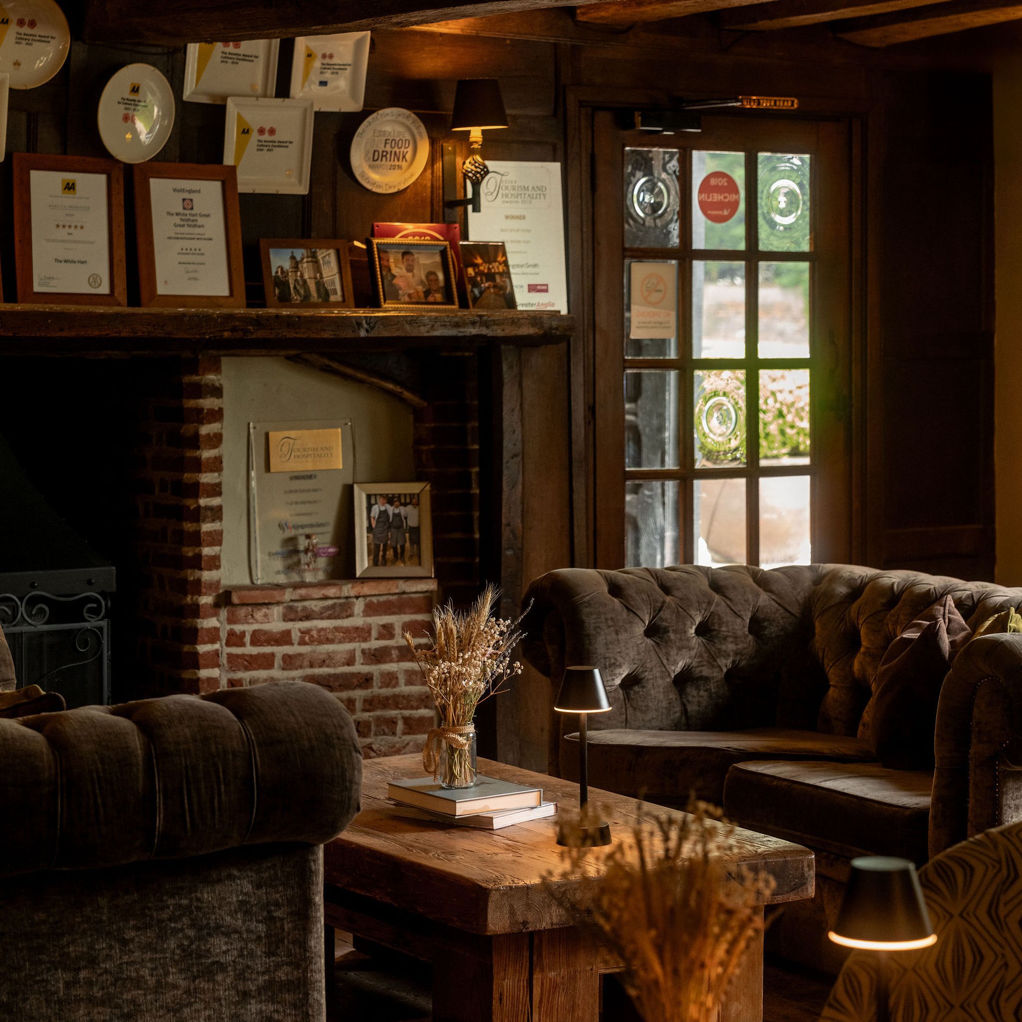 Cozy rustic lounge area with wooden beams, plush sofas, and a brick fireplace adorned with awards and framed pictures.